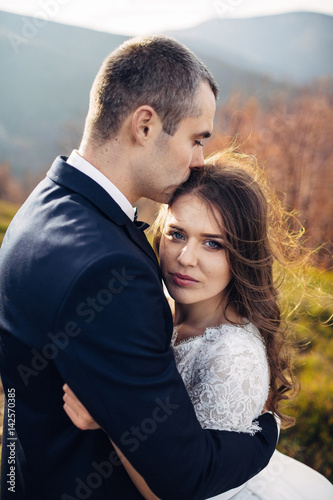Tall groom kisses bride's head while she leans to him on spring hill © myronovychoksana