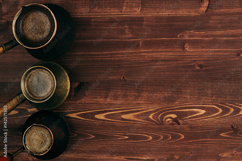 Hot coffee in shabby turkish pots cezve with tasty crema with copy space on brown old wooden board background, top view. Rustic style. Hot coffee in shabby turkish pots cezve with tasty crema with copy space on brown old wooden board background, top view. Rustic style.