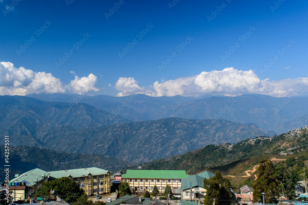 A hilltop view of house' roof with mountains and blue sky with clouds from Darjeeling Himalayan railway station on a misty morning.
