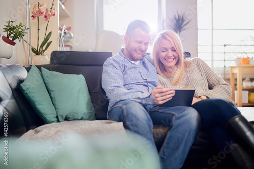 Caucasian happy couple using tablet computer while sitting on sofa in living room