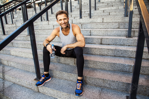 Happy smily sportsman resting and holding bottle of water, sitting on steps looking at camera and smiling . fitness, sport, exercising and people healthy lifestyle concept.