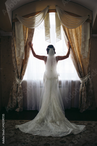 The bride in the dress is standing by the window