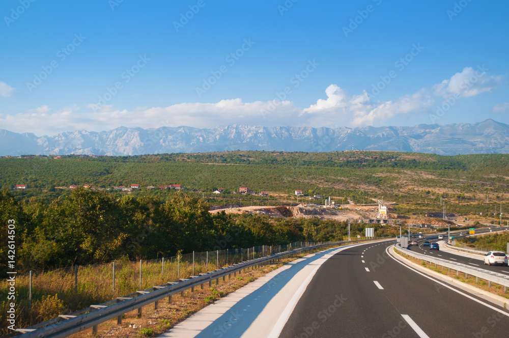 a highway in Croatia against the mountain range, many green trees and little houses on a warm summer day. Blue sky with several white clouds on the back