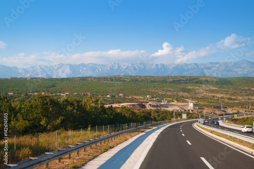 a highway in Croatia against the mountain range  many green trees and little houses on a warm summer day. Blue sky with several white clouds on the back
