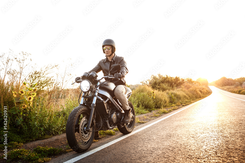 Fototapeta premium Młody przystojny mężczyzna, jazda na motocyklu w wiejskiej drodze. Tło światło słoneczne pochodni.