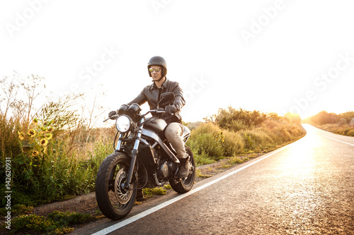 Young handsome man riding on motorbike at countryside road. Flare sunlight background.