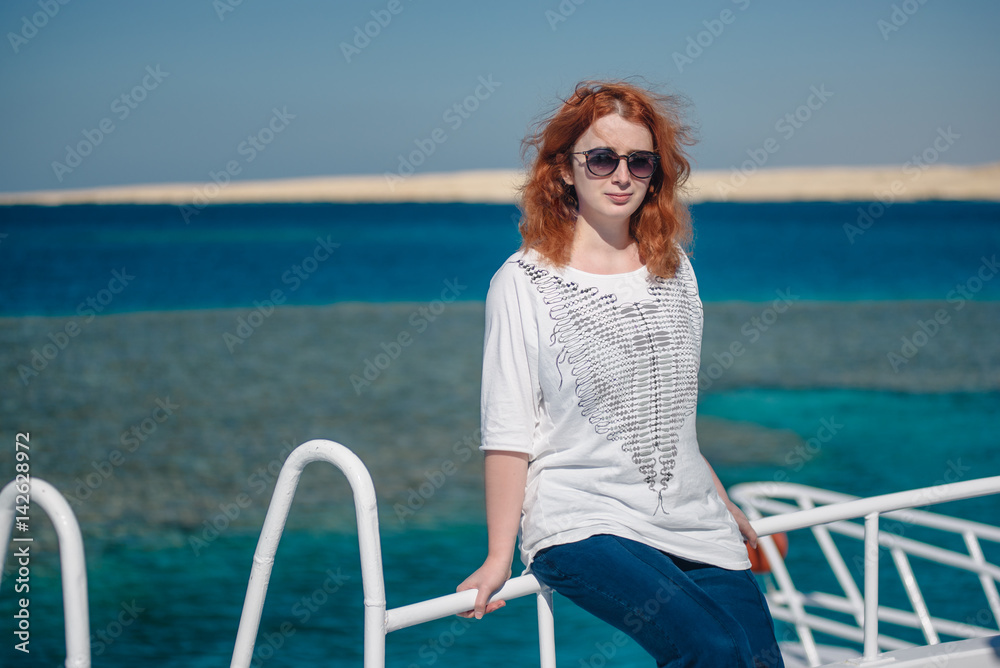 Beautiful ginger woman in sun glasses sits on a white yacht in a sea with clear turquoise water. Relaxation at summer vacation under a sun. Woman looks to the camera.