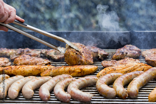 Fleisch und Wurst werden auf einem Grill zubereitet und mit einer Zange gewendet