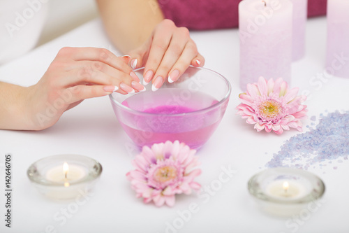 Spa Procedure. Woman In Beauty Salon Holding Fingers In Aroma Bath For Hands. Closeup Of Female Nails Soaking In Bowl Of Water With Floating Pink Flower Petals. Skin Care. High Resolution