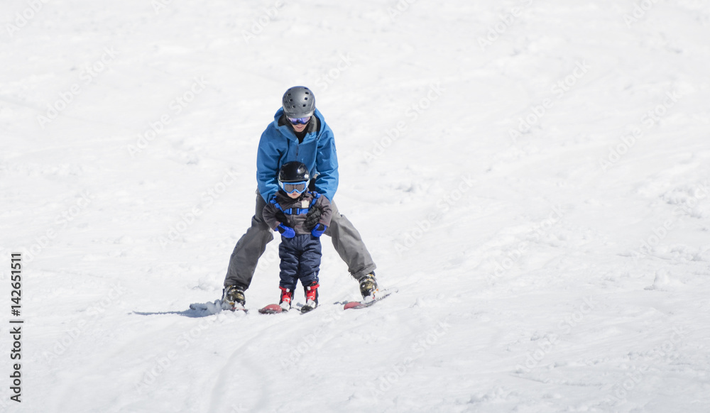 Toddler Boy Skis Downhill with the Help of His Father. Dressed Safely with Helmet and Harness.
