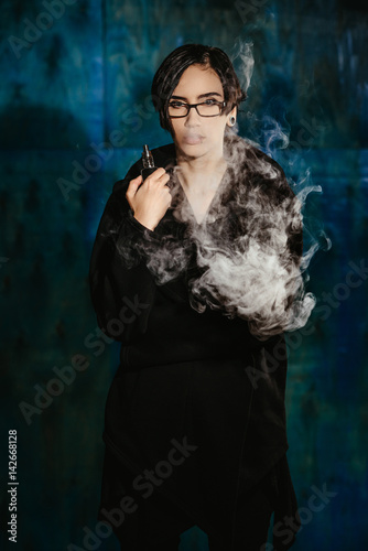 A girl with glasses vaping and releases a cloud of vapor. Model in a black vaper smoke vaporizer on a turquoise background