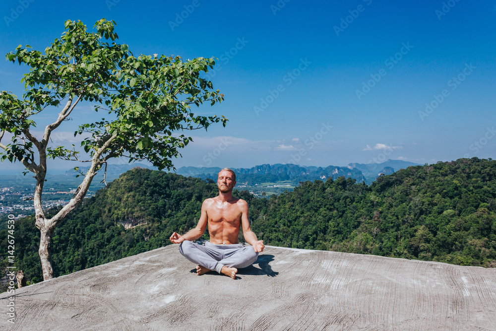 Fit man meditating on nature. Young shirtless male sitting on ground in lotus pose with eyes closed on background of tropic valley.