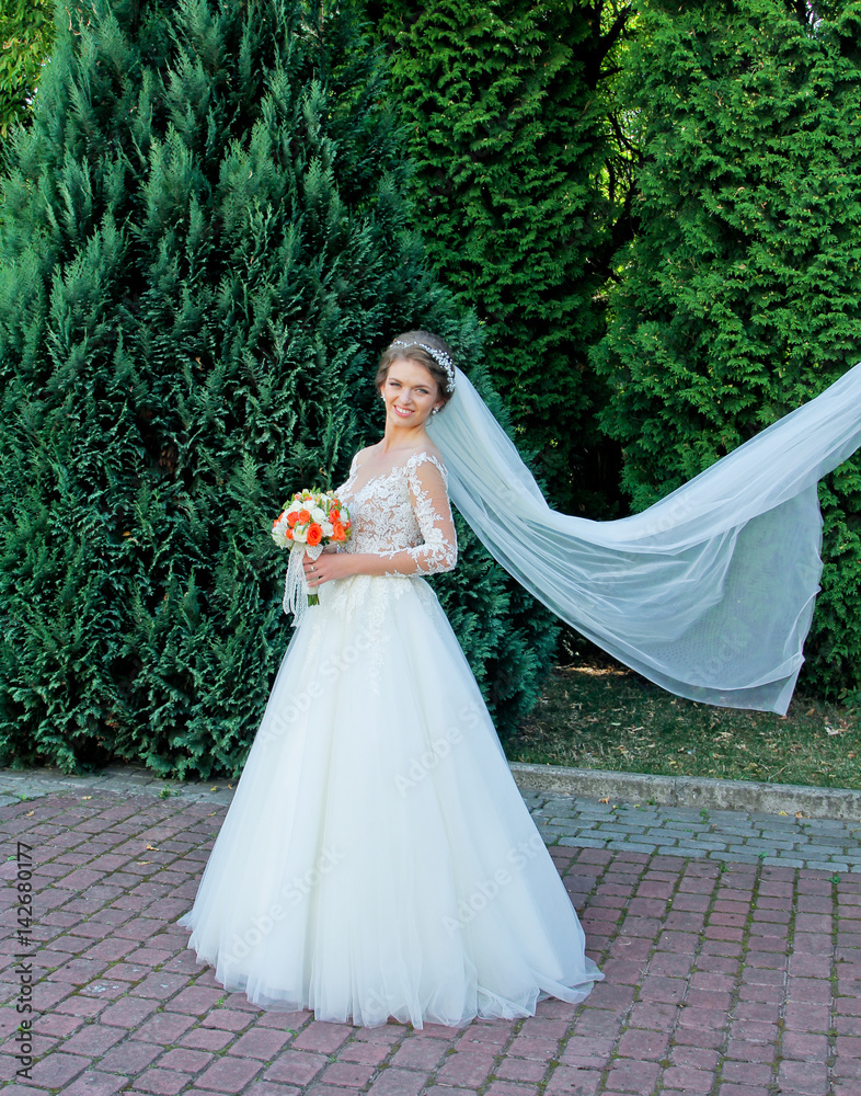 Beautiful light brown bride play with veil on the wind. Attractive girl standing in the park on their wedding day