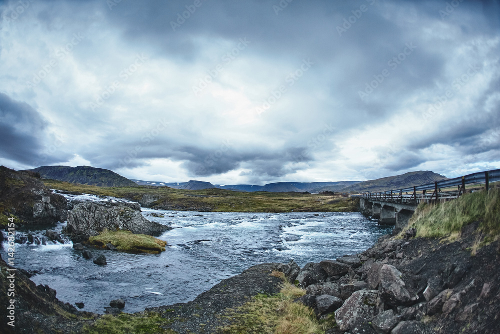 Mountain river in the severe gloomy Icelandic landscape. Stones and moss in the foreground. The picture was taken in cloudy cloudy weather in the autumn afternoon. The picture shows a bridge across