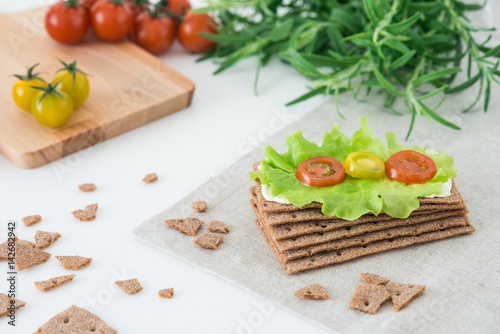 Rye crispy bread (Swedish crackers) with soft cheese, lettuce leave, cherry tomatoes and rosemary on white background. Healthy snack concept