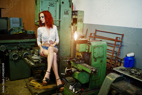 Red haired girl wear on short denim shorts and white blouse posed at industrial machine at the factory. photo