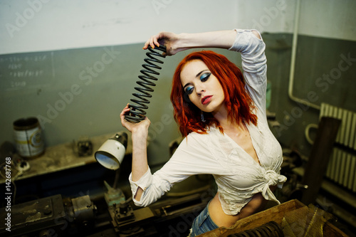 Red haired girl wear on short denim shorts and white blouse with steel spring at hand posed at industrial machine at the factory. photo