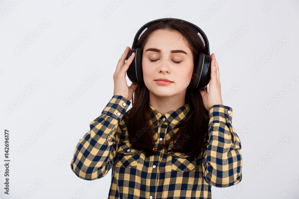 Young beautiful brunette girl dressed in plaid shirt enjoing music in headphones with eyes closed over white background Young beautiful brunette girl dressed in plaid shirt enjoing music in headphones with eyes closed over white background