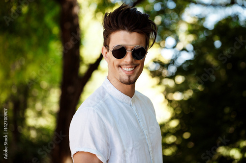 Young successful businessman in sunglasses smiling, walking around sunny city.