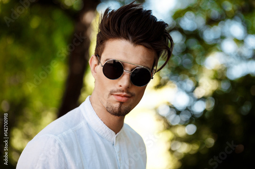 Young successful businessman in sunglasses smiling, walking around sunny city.