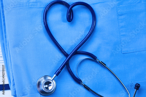 Medical stethoscope twisted in heart shape lying on patient medical history list and blue doctor uniform closeup. Medical help or insurance concept. Cardiology care, health, protection and prevention