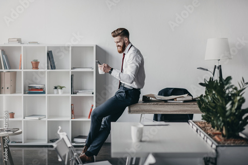 young businessman using smartphone and drinking coffee in office