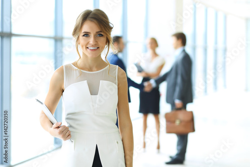Free Download Blonde woman with touchpad computer looking at camera and smiling while business people are shaking hands over office background - GFXLoad