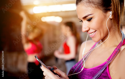 Young attractive fitness model listening music on smart phone charching positive energy before workout outside. Sun is shining and friends are behind preparing for training.