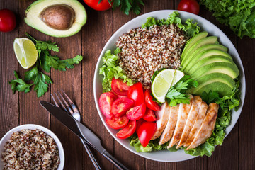 Healthy salad bowl with quinoa, tomatoes, chicken, avocado, lime and mixed greens (lettuce, parsley) on wooden background top view. Food and health.