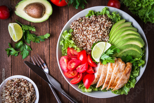 Healthy salad bowl with quinoa, tomatoes, chicken, avocado, lime and mixed greens (lettuce, parsley) on wooden background top view. Food and health.