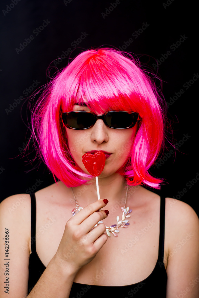 Fototapeta premium Beauty Woman With Pink Wig And Sunglasses and holding with her hand a candy with heart shape