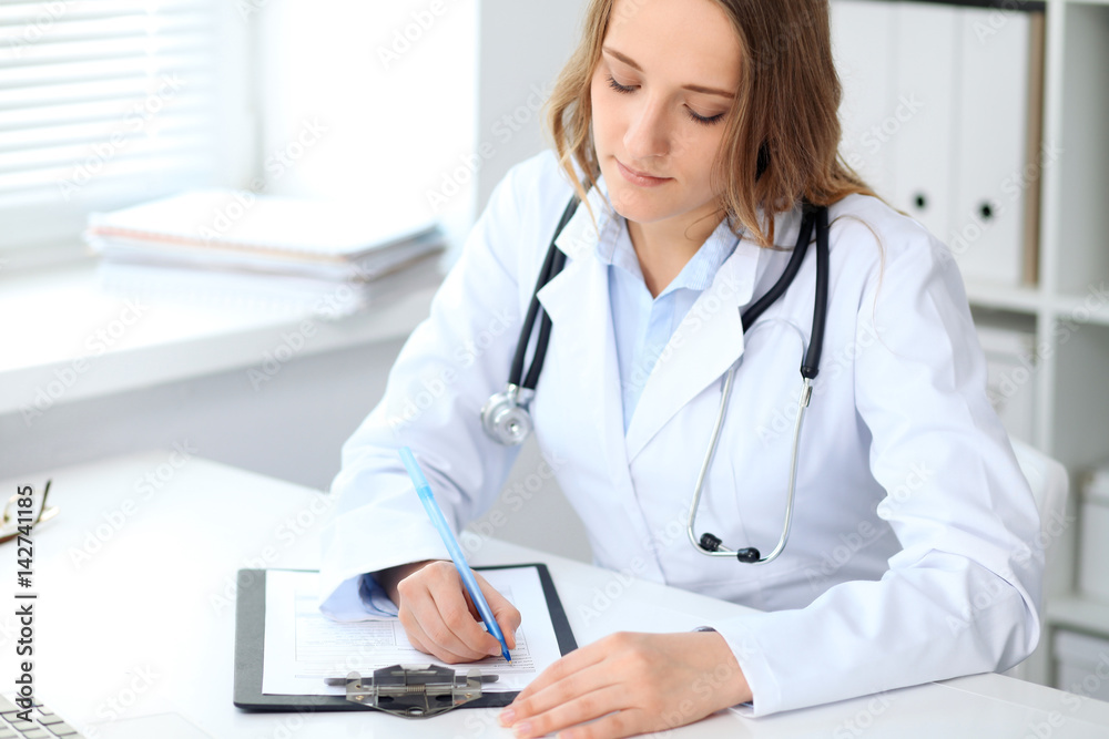 Young female doctor filling up medical form while sitting at the table near window in hospital Young female doctor filling up medical form while sitting at the table near window in hospital