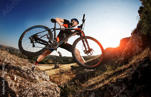 Professional Cyclist Taking his Bike up the Rocky Trail at evening. Extreme Sport Concept. Low angle and fisheye.