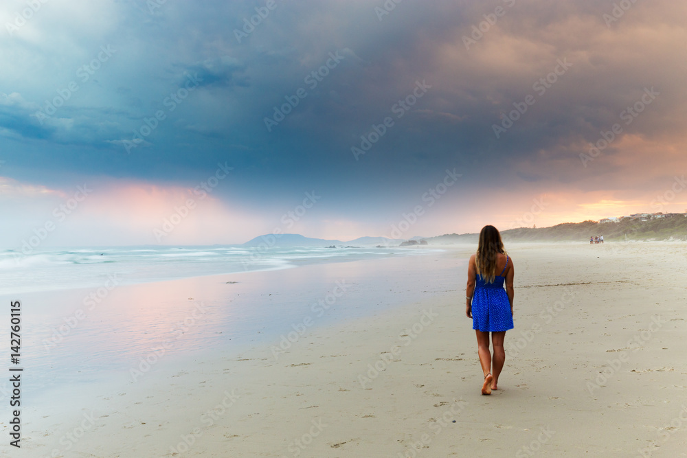 A young woman walks on an empty Australian beach under beautiful illuminated storm clouds.