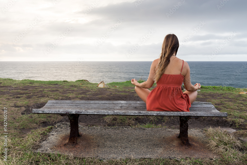 A woman in an orange dress meditates on a wooden bench by the sea.