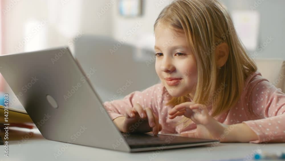 Cute Young Girl Uses Laptop Computer with Interest while Sitting at the Table in Her Light Room. Shot on RED EPIC-W 8K Helium Cinema Camera. Cute Young Girl Uses Laptop Computer with Interest while Sitting at the Table in Her Light Room. Shot on RED EPIC-W 8K Helium Cinema Camera.