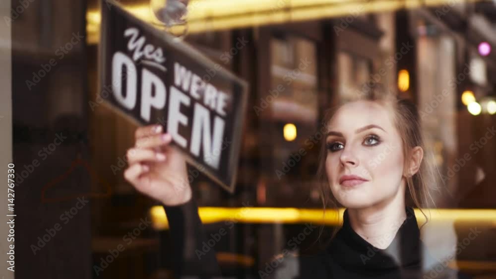 Attractive young blonde woman owner turns sign from close to open and shares a friendly, bright smile to camera. Inside the shop. Being friendly. Successful morning, having a break.