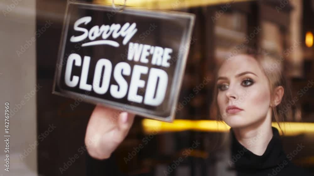 Beautiful waitress comes to the café entrance and flips the open sign to close, then looks around and goes away into the shop/café. The end of the working day. Working hard, being tired.