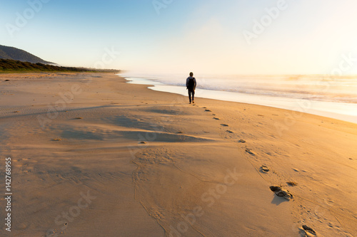 A person walking on a beautiful beach leaves a trail of footprints as the sunrise illuminates mist rising from the sea in golden light.