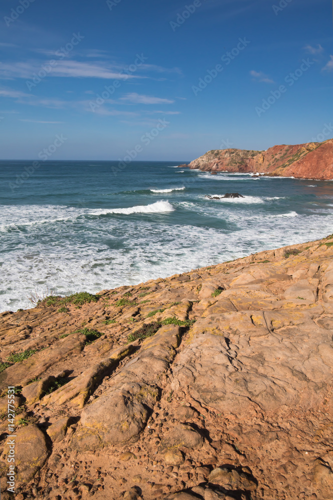 red soil rocks on colorful amado beach with waves on atlantic coastline, algarve, portugal