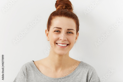Headshot of relaxed young ginger female hipster with perfect beautiful smile looking with happy expression at camera, rejoicing at her carefree lifestyle, posing indoors against white wall background