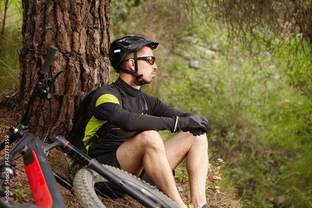 Cyclist sitting on ground at big tree in forest near his motor-powered electric bicycle waiting for friends before going mountain biking together on Sunday morning, enjoying fresh air and wild nature