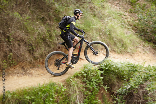 Young Caucasian professional rider performing mountain biking stunts on two-wheeled motor-powered bicycle on trail along cliff using pedal-assist system. Male biker cycling outdoors on electric bike
