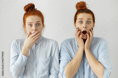 Human emotions and feelings. Face expressions. Two red haired astonished Caucasian students looking like twins with hair knots dressed in shirts. Bug-eyed sisters learned shocking information