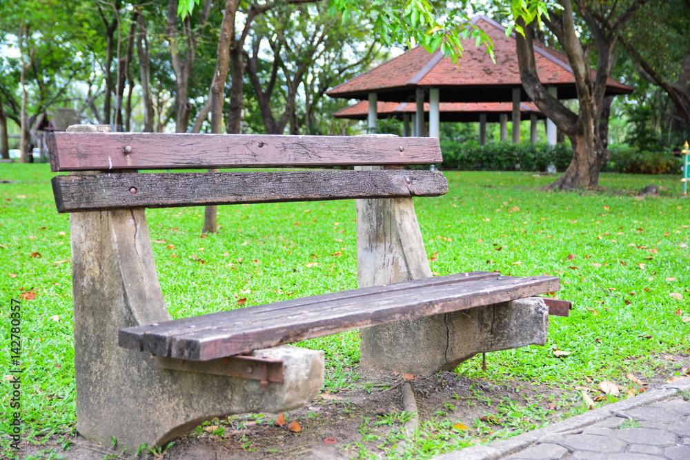 Wooden bench in public park with greenery grass field as background and red roof pavilion.