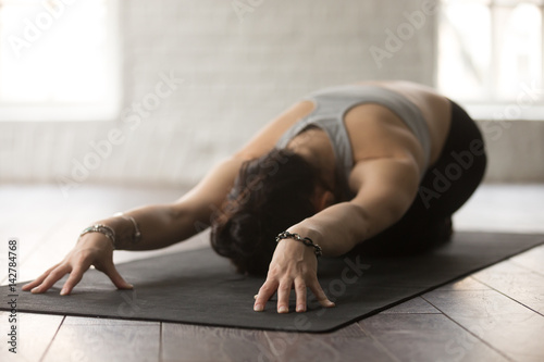 Young beautiful sporty yogi woman practicing yoga concept, sitting in Child exercise on black yogic mat, Balasana pose, working out, wearing sportswear, white studio or home background, wooden floor