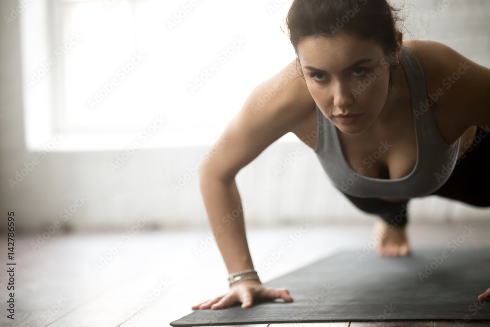 Fototapeta premium Young sporty cool woman practicing yoga, doing Push ups or press ups exercise, working out on black mat wearing sportswear, close up portrait, loft background. Weight loss, healthy lifestyle concept