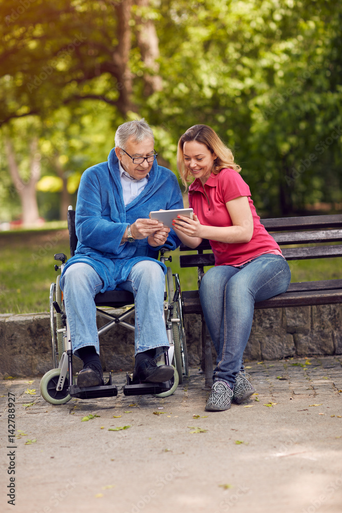 Disabled retiree man in park using tablet with daughter. Disabled retiree man in park using tablet with daughter.