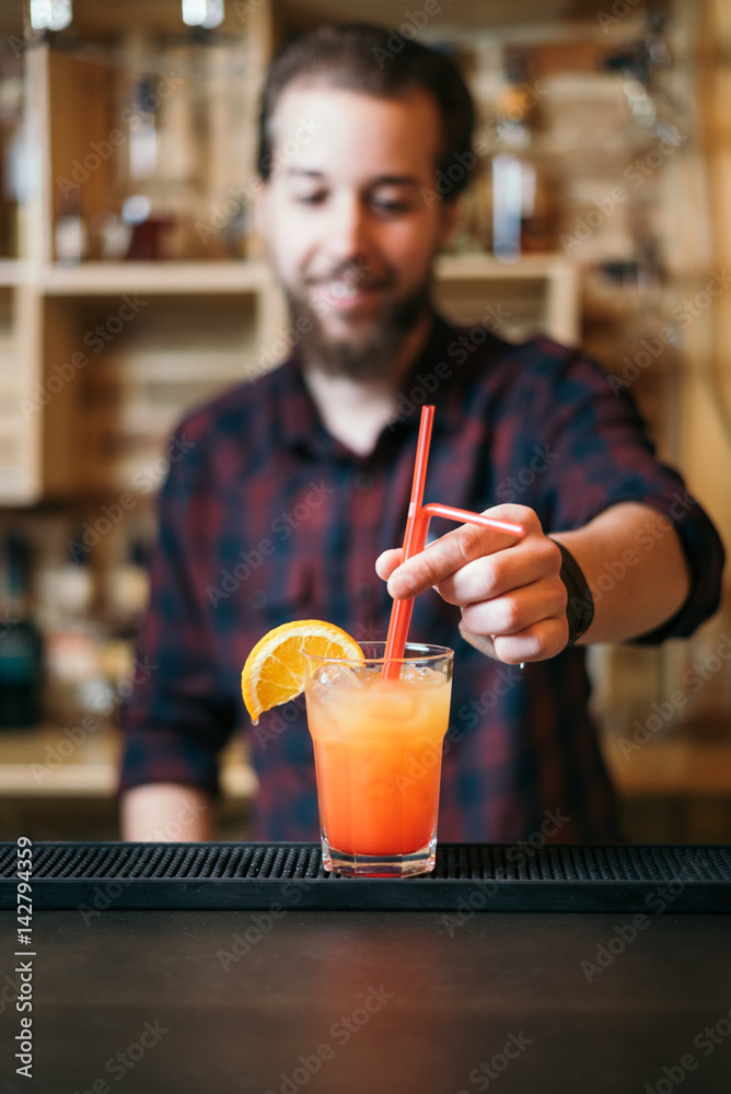 Fototapeta premium Handsome bearded barman is making tequila sunrise cocktail in night club. Selective focus on glass.
