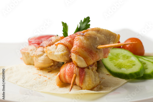 chicken on a skewer, baked in bacon, rests on a pita with cucumber, cherry tomatoes, parsley and savory sauce, white background, isolated